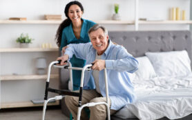 Female,Nurse,Helping,Elderly,Male,With,Walking,Frame,Stand,Up