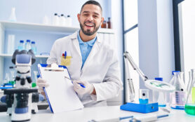 Hispanic,Man,Working,At,Scientist,Laboratory,Holding,Blank,Clipboard,Smiling